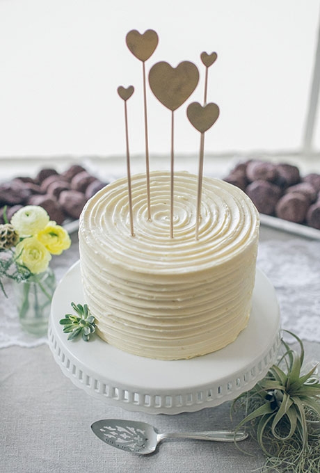 A simple one-tier white wedding cake with heart toppers created by Cake Monkey Bakery.